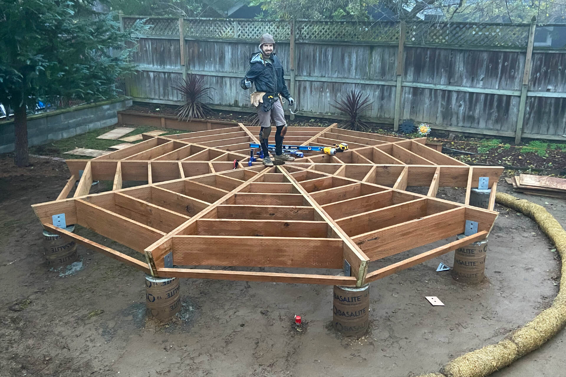 A construction photo of the builder standing alone atop the floor framing, looking at the camera and giving a thumbs-up.